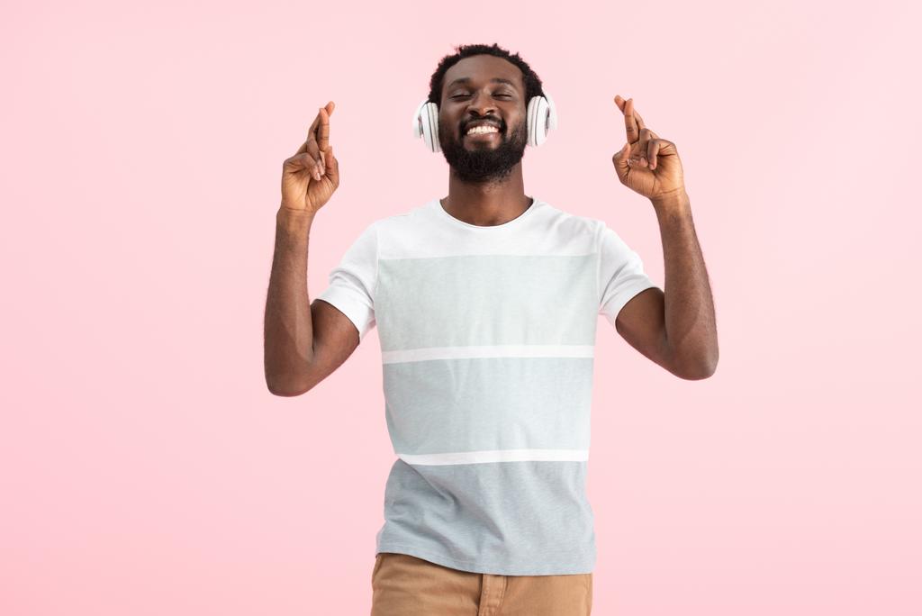 šťastný afroamerický muž s zavřenýma očima a překřížené prsty naslouchající hudbě se sluchátky, izolované na růžovém - Fotografie, Obrázek
