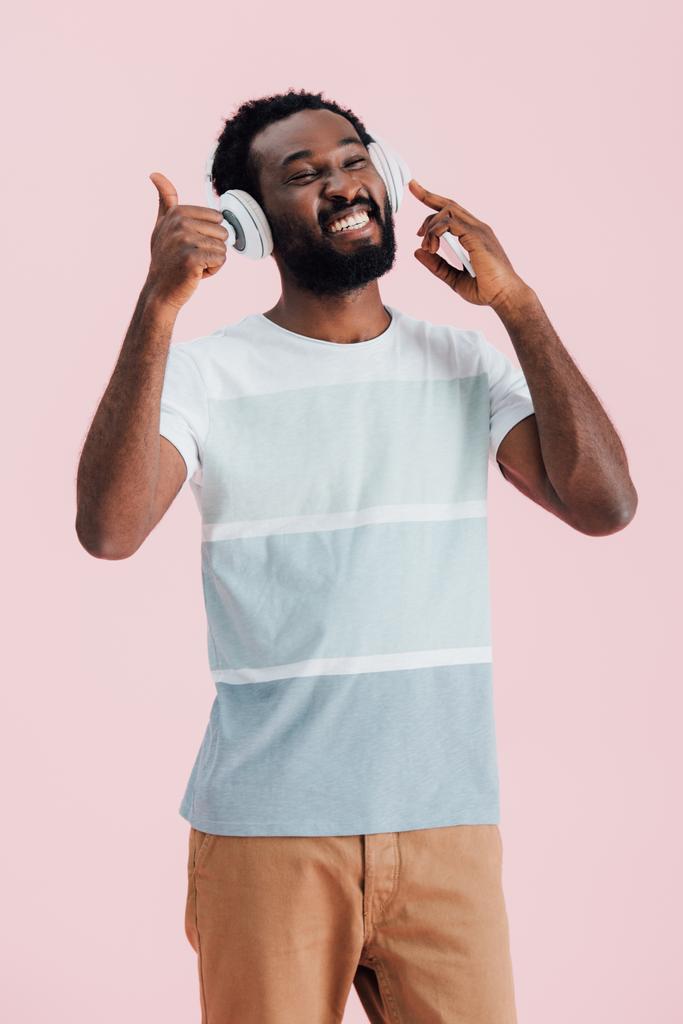 Wesoła African American Man słuchania muzyki ze słuchawkami i pokazując kciuka w górę, izolowane na różowy - Zdjęcie, obraz