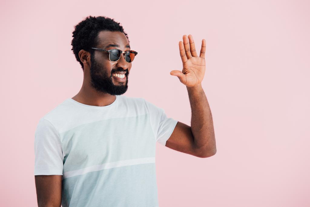 fröhliche junge afrikanisch-amerikanische Mann mit Sonnenbrille winkt isoliert auf rosa - Foto, Bild