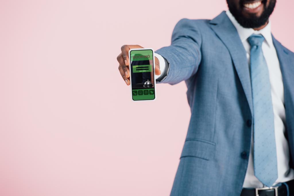 vista recortada del hombre de negocios afroamericano en traje que muestra el teléfono inteligente con aplicación de reserva, aislado en rosa
  - Foto, imagen