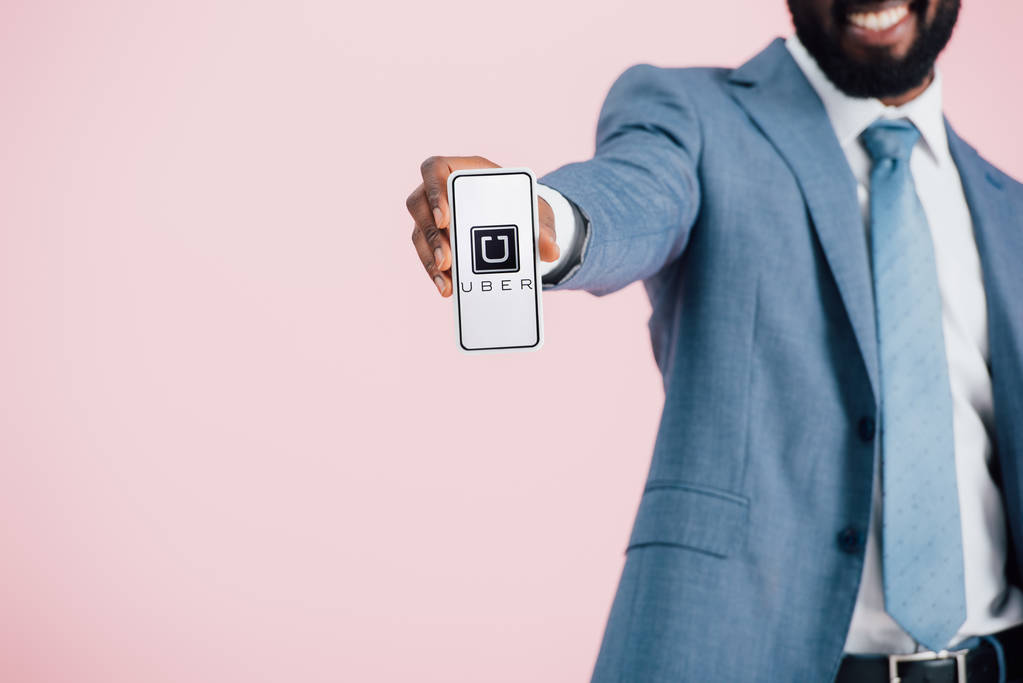 Київ, Україна-17 травня 2019: обрізаний вид афроамериканського бізнесмена в костюмі показує смартфон з додатком Uber, ізольований на рожевому  - Фото, зображення