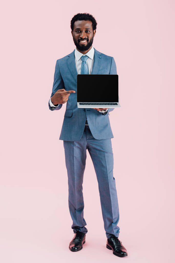 ピンクに隔離された空白の画面でラップトップを指す笑顔のアフリカ系アメリカ人ビジネスマン - 写真・画像