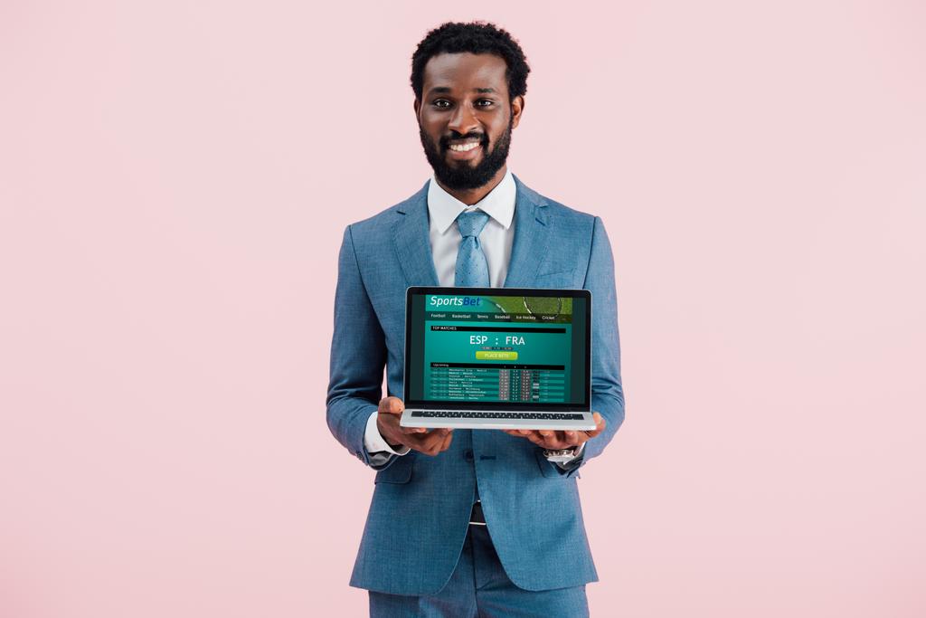 χαμογελαστός Αφρικανός Αμερικανός επιχειρηματίας δείχνοντας φορητό υπολογιστή με αθλητικό στοίχημα ιστοσελίδα απομονωμένη στο μπλε - Φωτογραφία, εικόνα