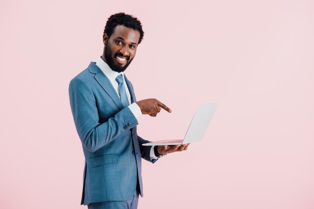 ピンクに隔離されたラップトップを指す陽気なアフリカ系アメリカ人ビジネスマン - 写真・画像