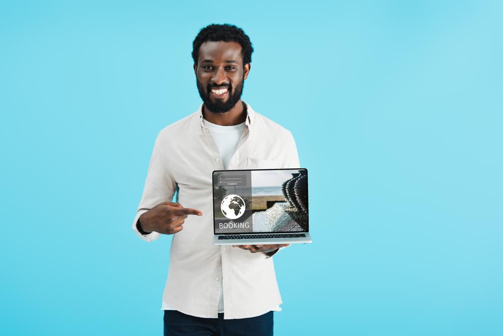 usmívající se Africká Američanka ukazující na notebook s rezervací webových stránek izolované na modré - Fotografie, Obrázek