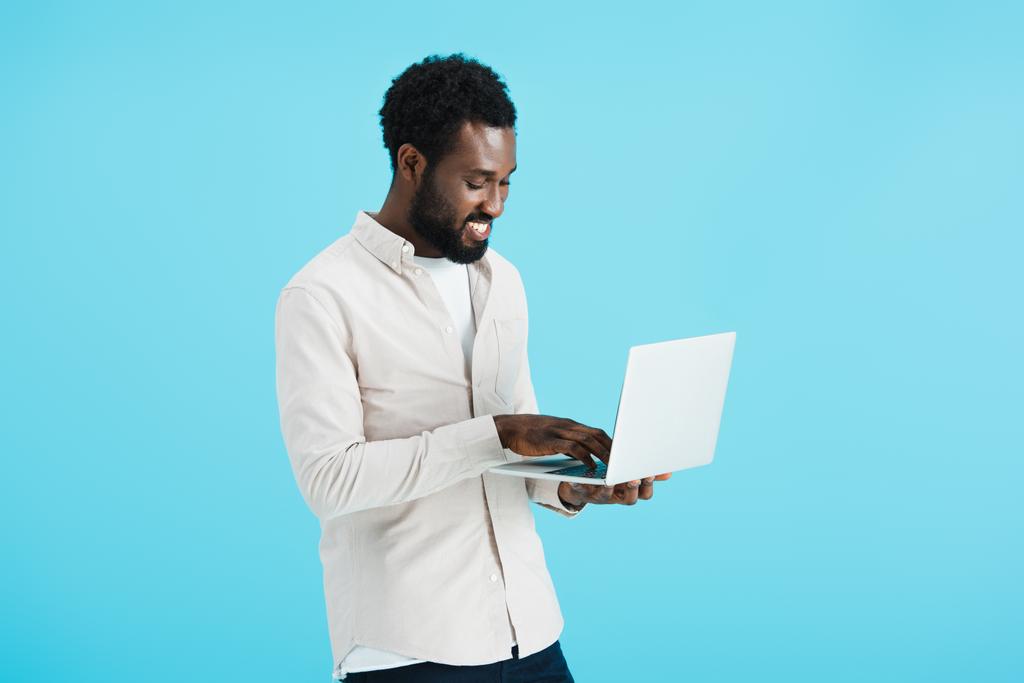 glimlachend African American man in wit shirt met behulp van laptop geïsoleerd op blauw - Foto, afbeelding