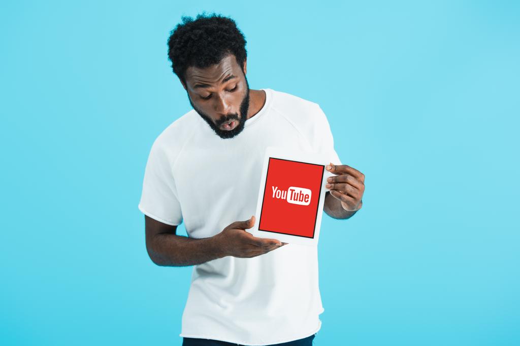 KYIV, UCRANIA - 17 de mayo de 2019: sorprendido hombre afroamericano mirando la tableta digital con la aplicación de youtube, aislado en azul
 - Foto, imagen