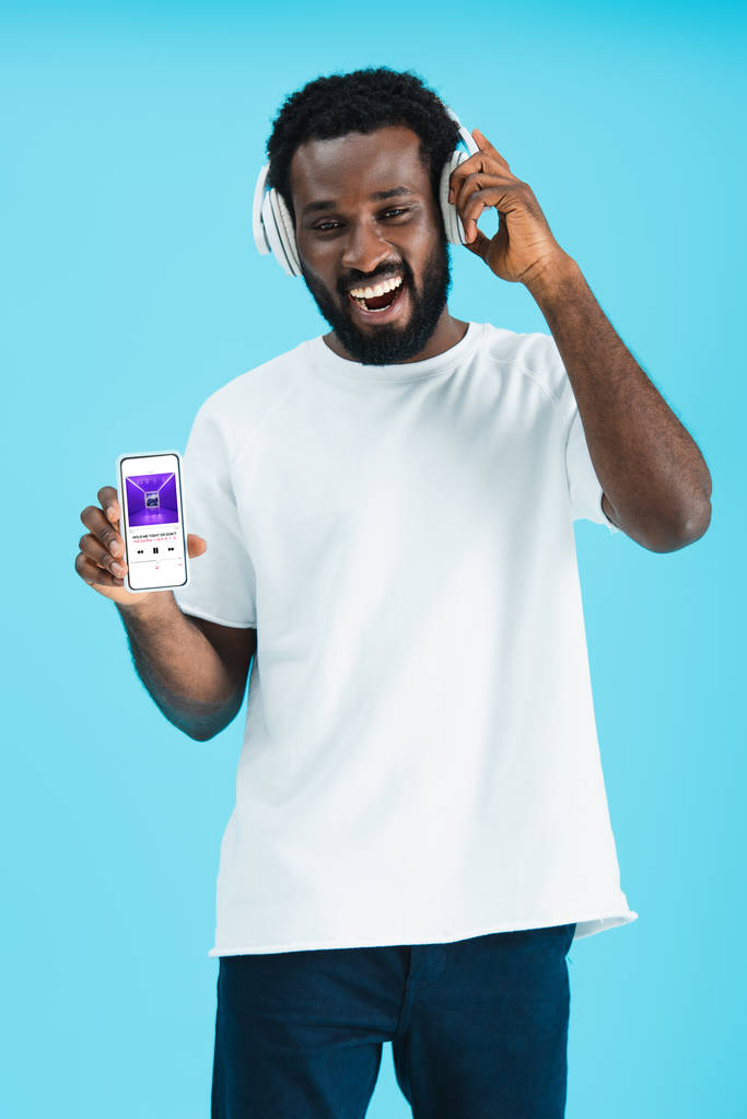 KYIV, UCRANIA - 17 DE MAYO DE 2019: hombre afroamericano sonriente escuchando música con auriculares y mostrando teléfono inteligente con aplicación de música de manzana, aislado en azul
 - Foto, imagen