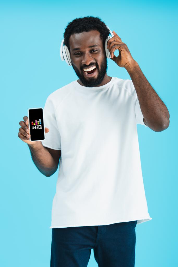 КИЕВ, УКРАИНА - 17 мая 2019 года: улыбающийся африканский американец слушает музыку в наушниках и показывает смартфон с приложением Deezer, изолированным на синем
 - Фото, изображение