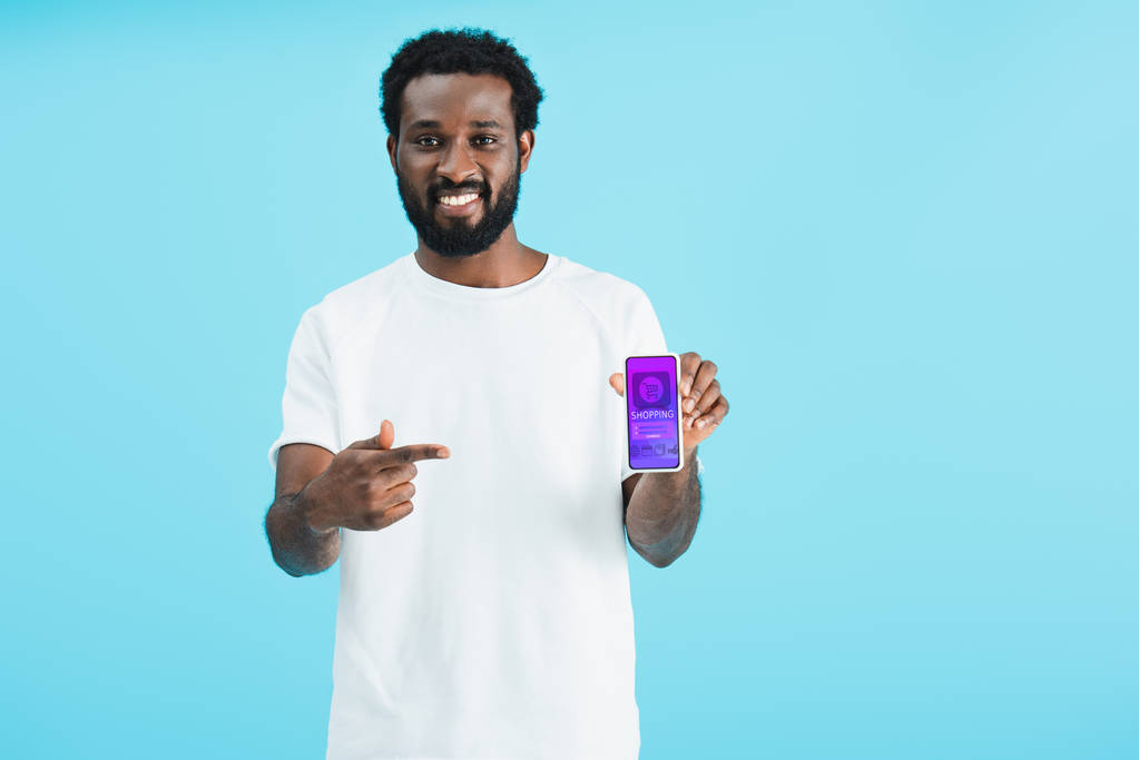 smiling african american man pointing at smartphone with shopping app, isolated on blue - Photo, Image