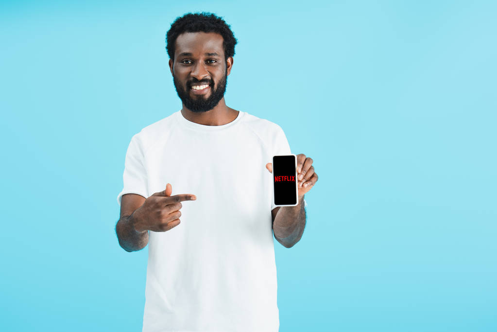Κίεβο, Ουκρανία-17 Μαΐου, 2019: χαμογελαστός Αφρικανός Αμερικανός άνθρωπος που δείχνει στο smartphone με την εφαρμογή Netflix, απομονώνεται σε μπλε - Φωτογραφία, εικόνα