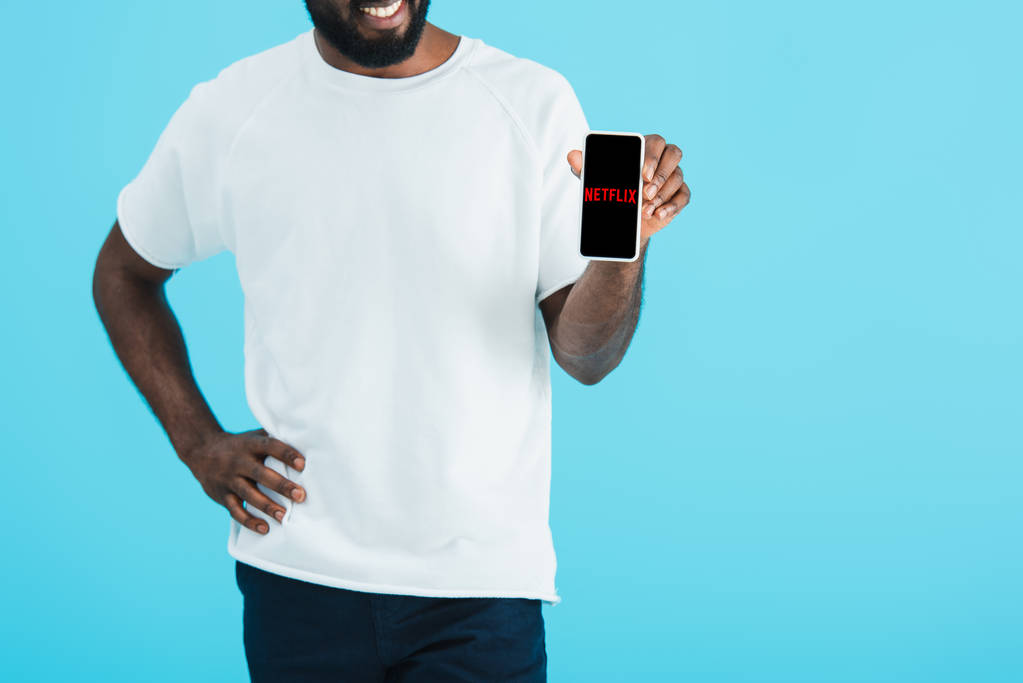キエフ、ウクライナ - 2019年5月17日:ネットフリックスアプリでスマートフォンを示すアフリカ系アメリカ人男性のトリミングビュー、青色で隔離 - 写真・画像