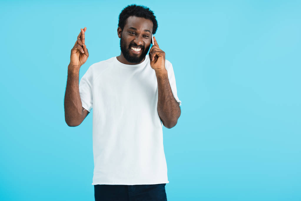 χαμογελαστός Αφρικανός Αμερικανός άντρας με σταυρωτά δάχτυλα που μιλούν στο smartphone, απομονωμένα στο μπλε - Φωτογραφία, εικόνα