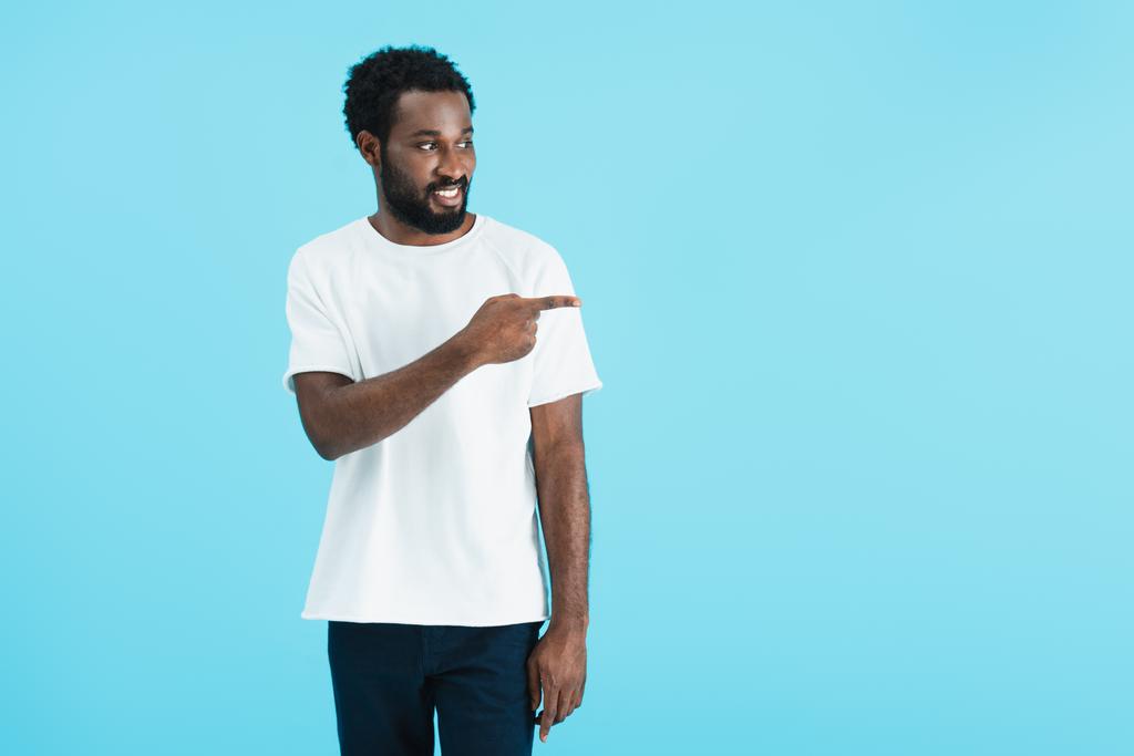 lächelnder afrikanisch-amerikanischer Mann in weißem T-Shirt, der vereinzelt auf blauem Grund zur Seite zeigt   - Foto, Bild