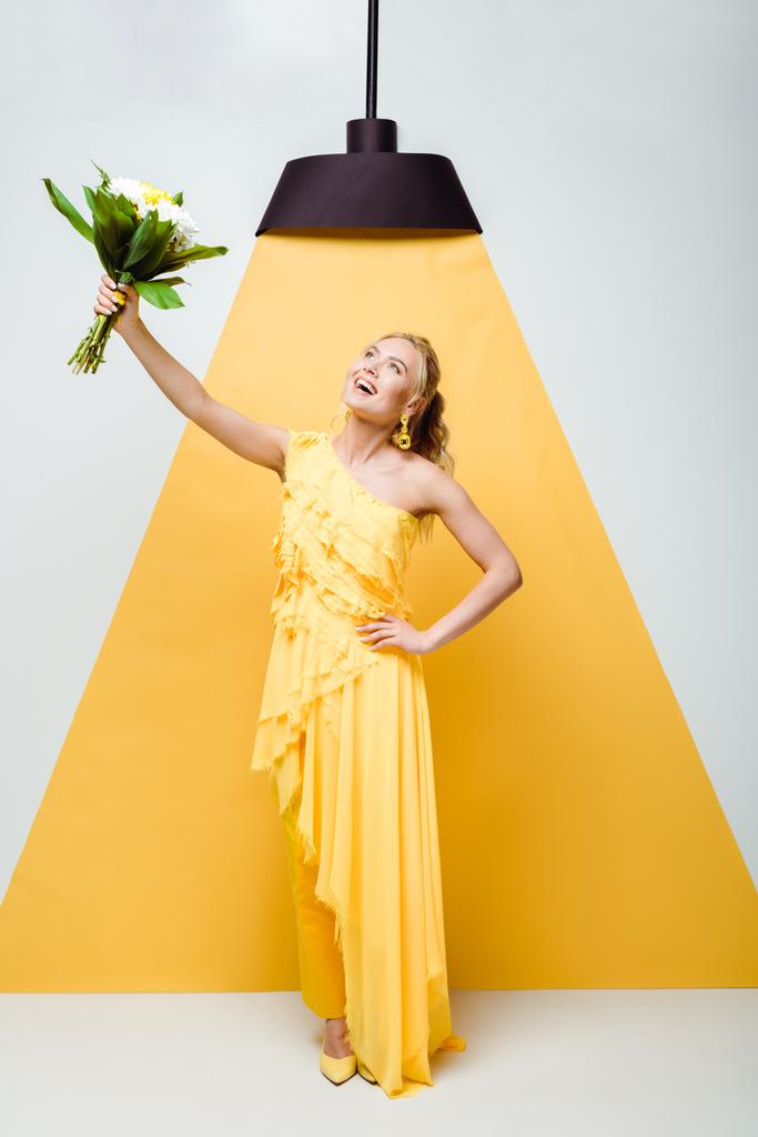 jovem mulher atraente de pé com a mão no quadril e segurando buquê de flores em branco e amarelo
  - Foto, Imagem