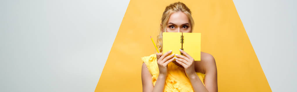 Panoramaaufnahme einer blonden jungen Frau, die ihr Gesicht mit einem Notizbuch in weiß und gelb bedeckt - Foto, Bild