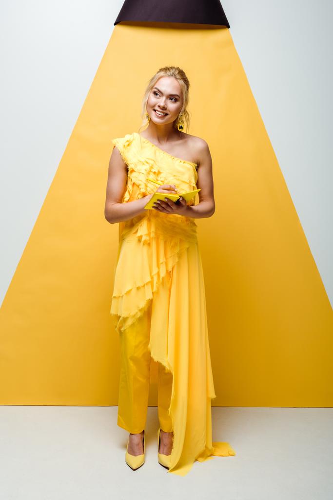 gelukkige jonge vrouw poseren en het houden van notebook met potlood op wit en geel  - Foto, afbeelding