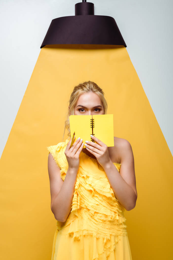 ξανθιά νεαρή γυναίκα που καλύπτει το πρόσωπο με το σημειωματάριο σε λευκό και κίτρινο - Φωτογραφία, εικόνα