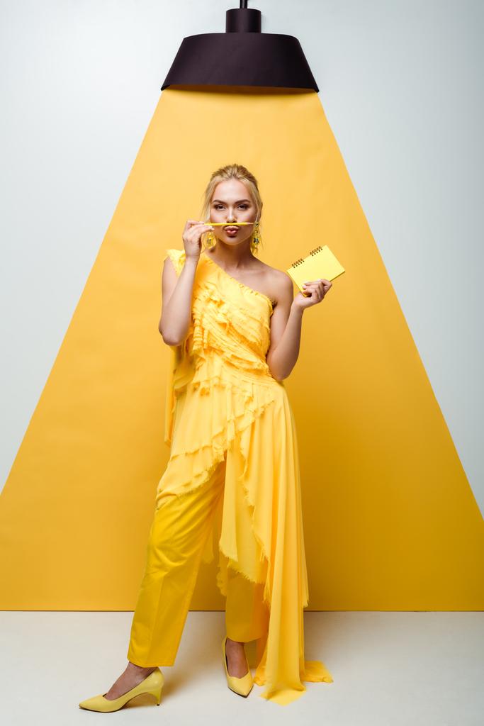 ξανθιά γυναίκα με πάπια πρόσωπο κρατώντας μολύβι κοντά στο στόμα και το σημειωματάριο στο χέρι σε λευκό και κίτρινο  - Φωτογραφία, εικόνα