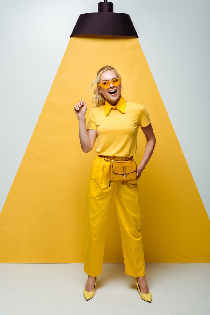 gelukkige blonde vrouw in zonnebril aanraken van haar terwijl poseren op wit en geel  - Foto, afbeelding