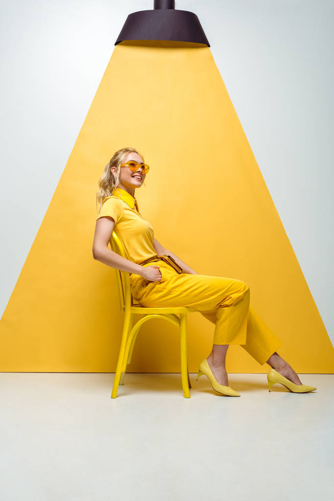 Gelukkig blonde vrouw in zonnebril zittend op stoel op wit en geel  - Foto, afbeelding
