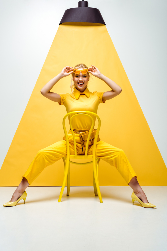 χαρούμενη ξανθιά γυναίκα αγγίζοντας τα γυαλιά ηλίου και κάθεται στην καρέκλα σε λευκό και κίτρινο  - Φωτογραφία, εικόνα