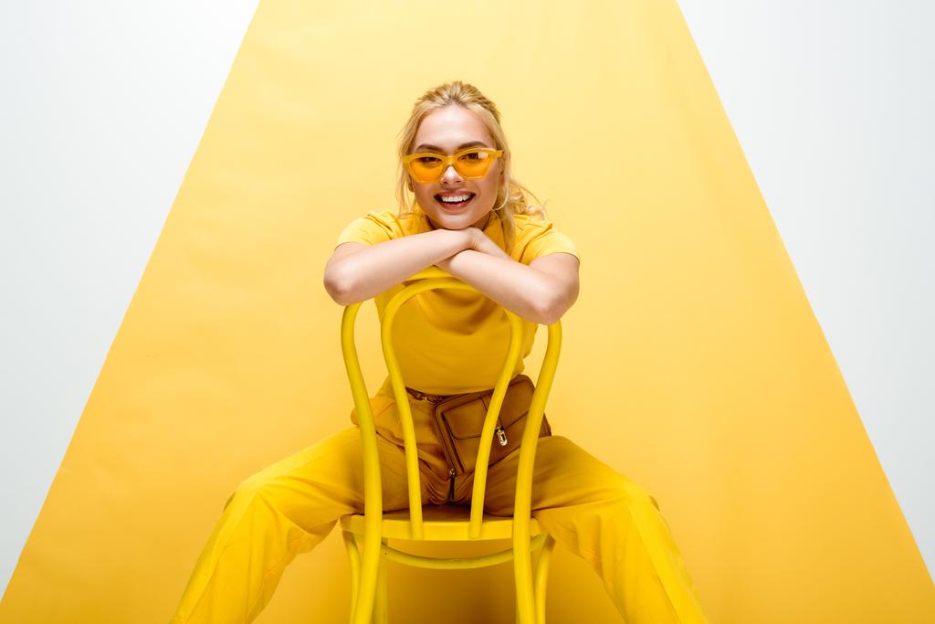 mulher loira alegre em óculos de sol sentado na cadeira em branco e amarelo
  - Foto, Imagem
