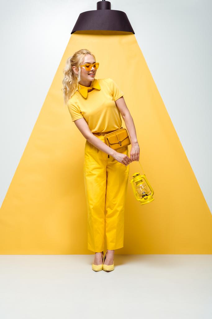 Gelukkig blonde vrouw in zonnebril poseren en het houden van vintage lamp op wit en geel  - Foto, afbeelding