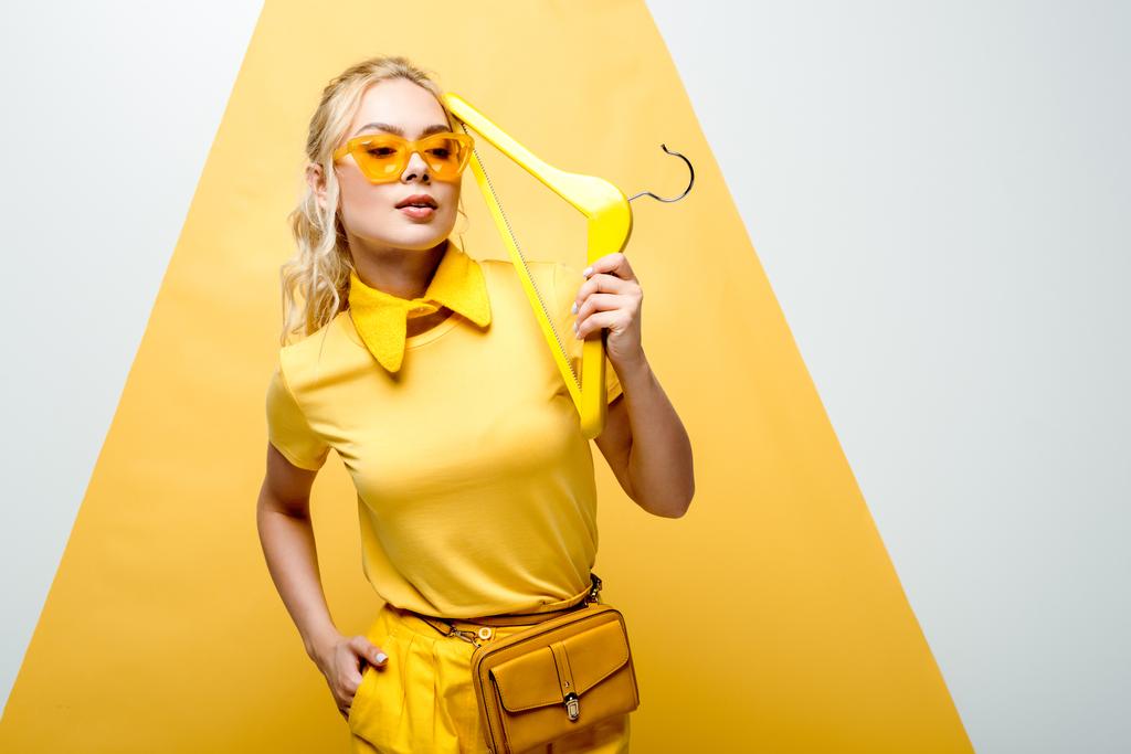 atraktivní blondýnka v brýlích s rukou v kapse a přidržením věšáku na bílém a žlutém  - Fotografie, Obrázek