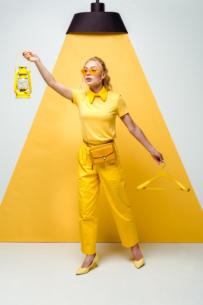 aantrekkelijke blonde vrouw in zonnebril houden hanger en retro lamp op wit en geel  - Foto, afbeelding