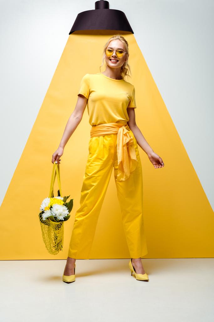 χαρούμενη ξανθιά γυναίκα κρατώντας τσάντα string με τα λουλούδια, ενώ ποζάρει σε λευκό και κίτρινο  - Φωτογραφία, εικόνα