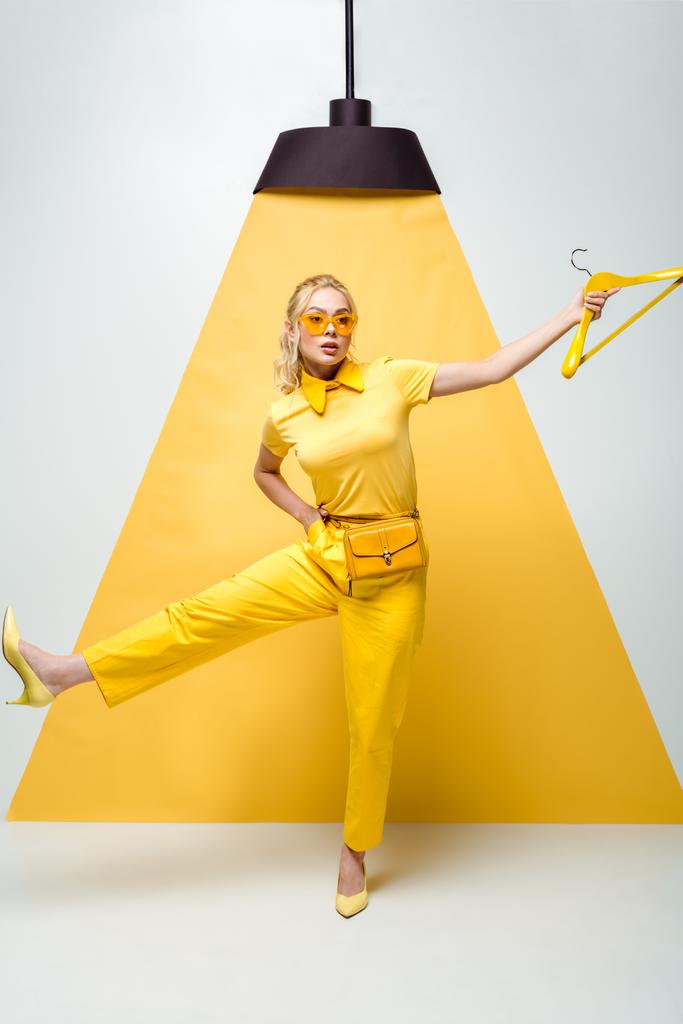 atrakcyjna blond kobieta w okulary trzymające wieszak i stwarzające na białym i żółtym  - Zdjęcie, obraz