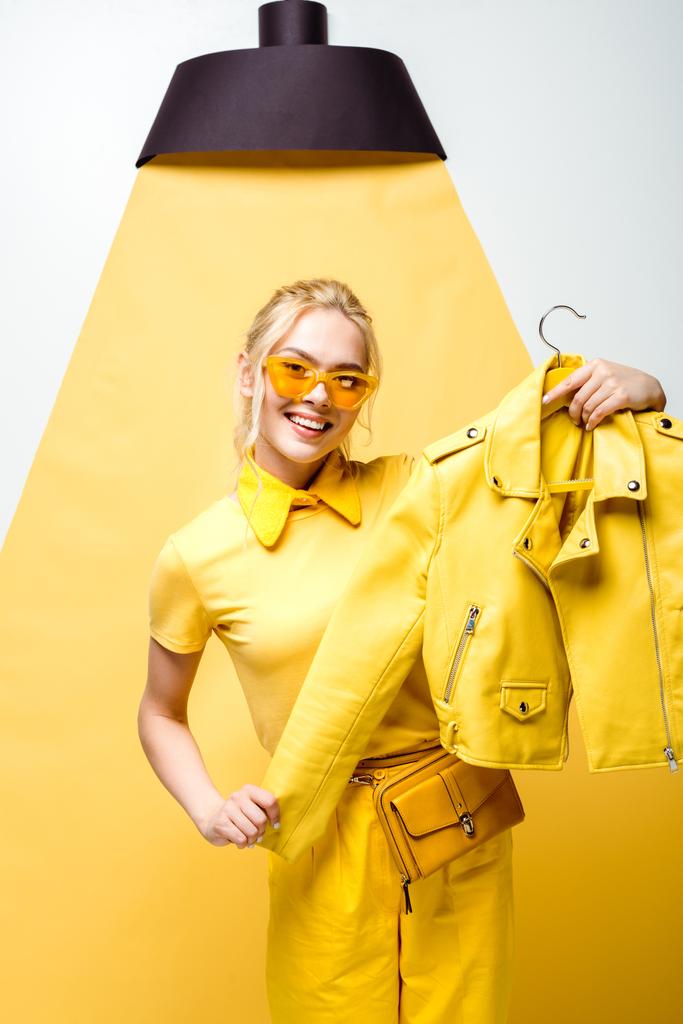šťastná blondýnka v brýlích s věšáku na bílém a žlutém plášti  - Fotografie, Obrázek