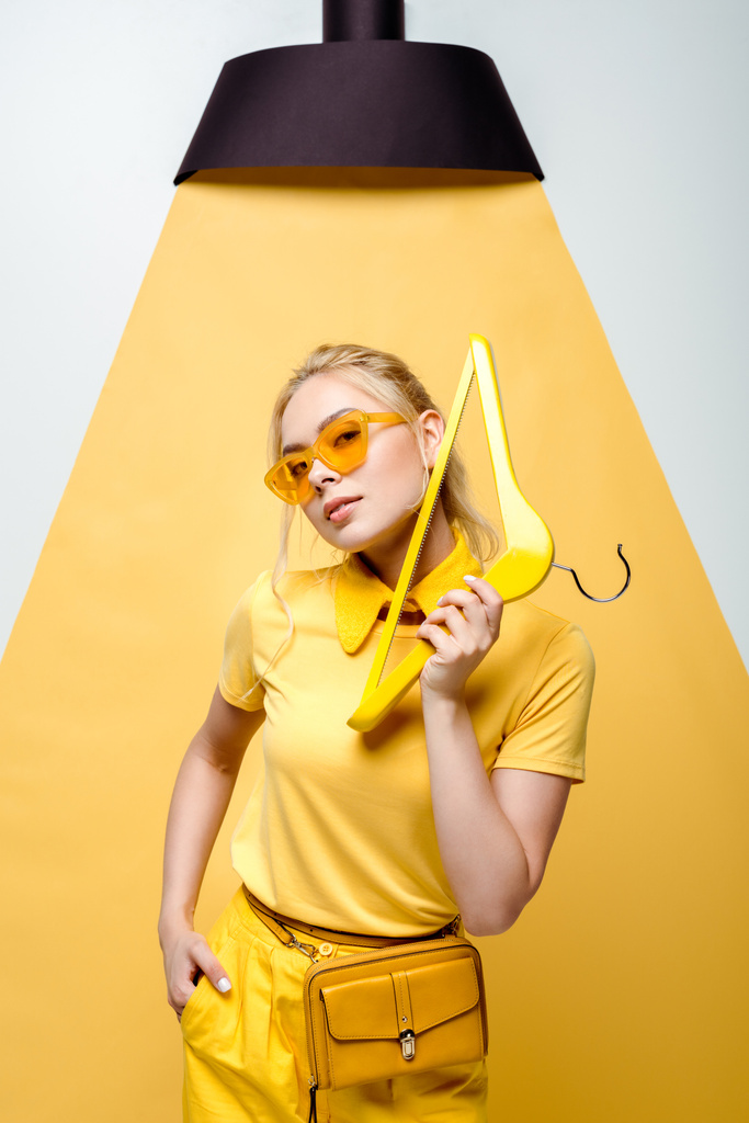 mulher loira atraente com a mão no bolso segurando cabide e posando em branco e amarelo
  - Foto, Imagem