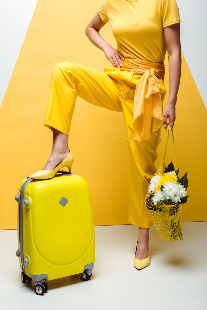 荷物の近くで腰に手を当ててポーズをとり、白と黄色の花で再利用可能なバッグを持つ女性のトリミングされたビュー  - 写真・画像