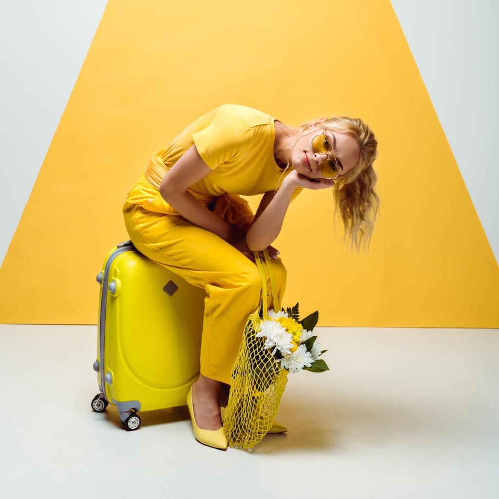 hübsches blondes Mädchen sitzt auf Gepäck und hält eine Tasche mit Blumen in weiß und gelb in der Hand  - Foto, Bild