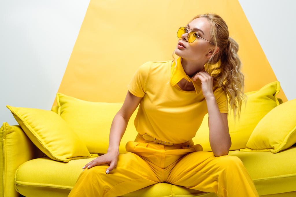 Elegantní blondýnka sedící na pohovce s polštáři na bílém a žlutém  - Fotografie, Obrázek