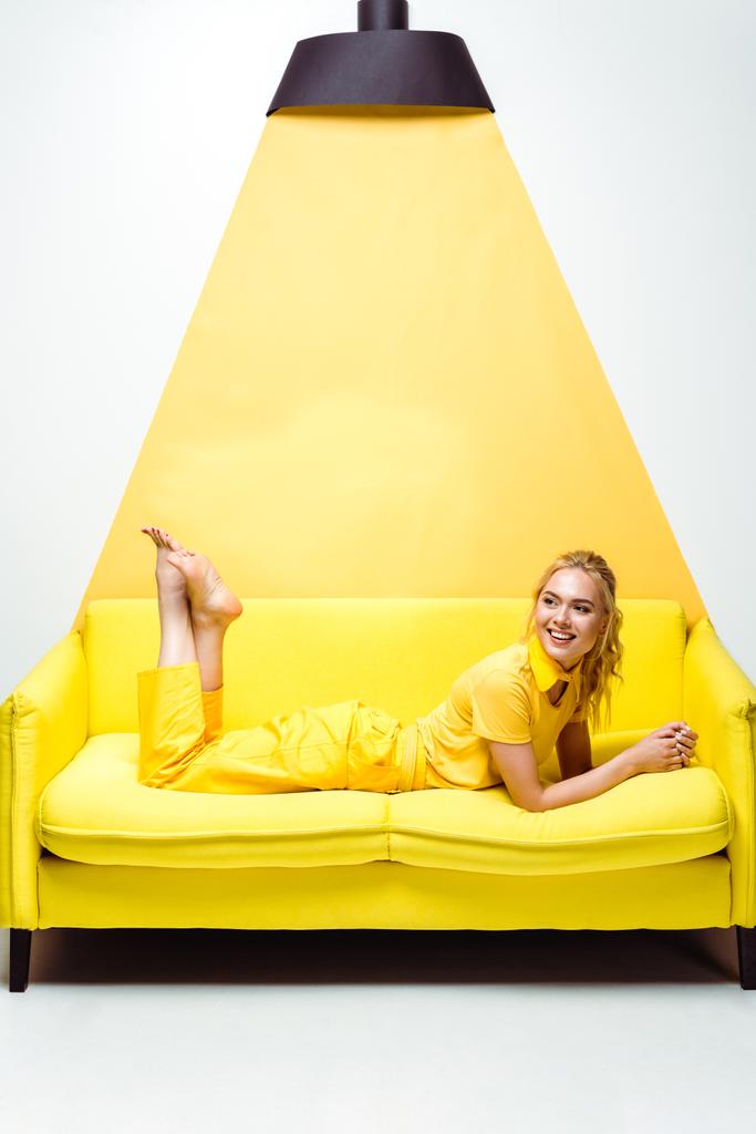 cheerful blonde girl with barefoot lying on sofa and smiling on white and yellow  - Photo, Image