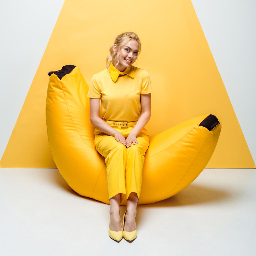 χαρούμενη ξανθιά γυναίκα που κάθεται στην καρέκλα τσάντα φασολιών σε λευκό και κίτρινο  - Φωτογραφία, εικόνα