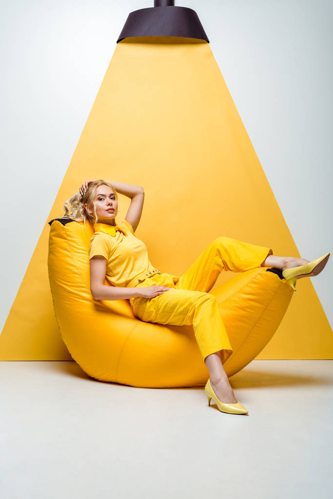 blonde girl sitting on bean bag chair and touching hair on white and yellow  - Photo, Image