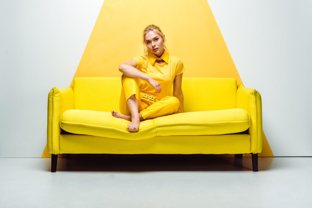 ξανθιά κορίτσι με ξυπόλητη κάθεται στον καναπέ και κοιτάζοντας την κάμερα σε λευκό και κίτρινο  - Φωτογραφία, εικόνα