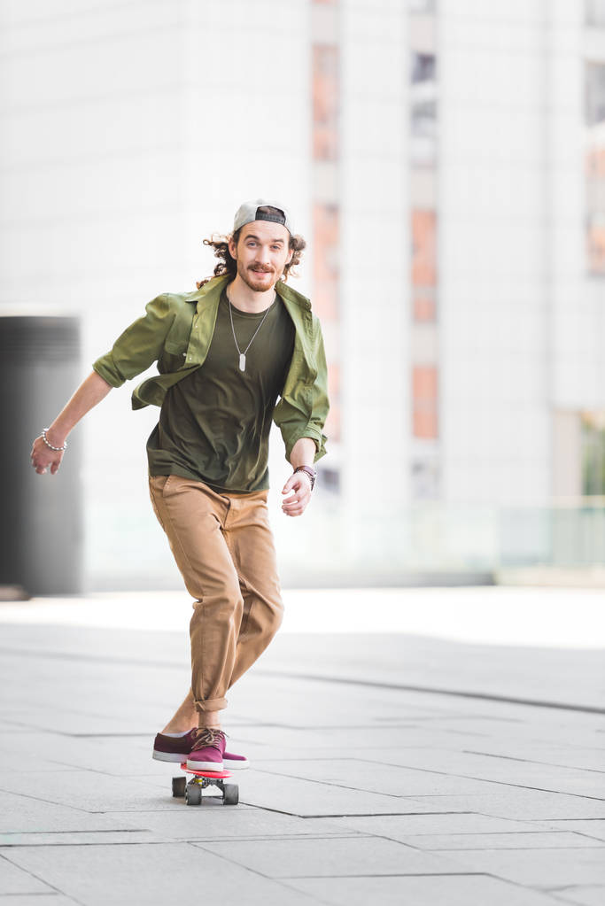 happy man looking at camera, riding on skateboard in city - Photo, Image