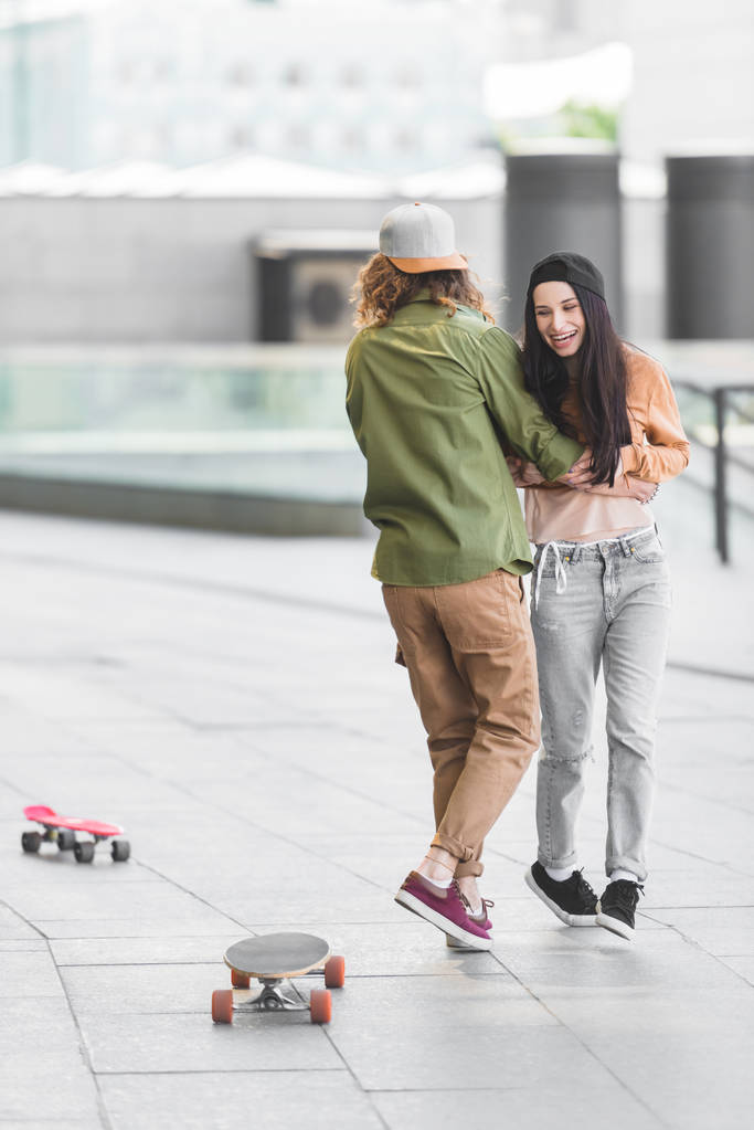 陽気な女性が男と抱き合い、市内のスケートボードの近くに立っている - 写真・画像