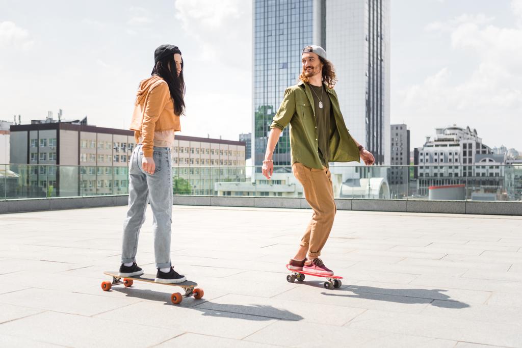 Mann in Freizeitkleidung schaut brünette Frau an, fährt auf Skateboard auf Dach - Foto, Bild