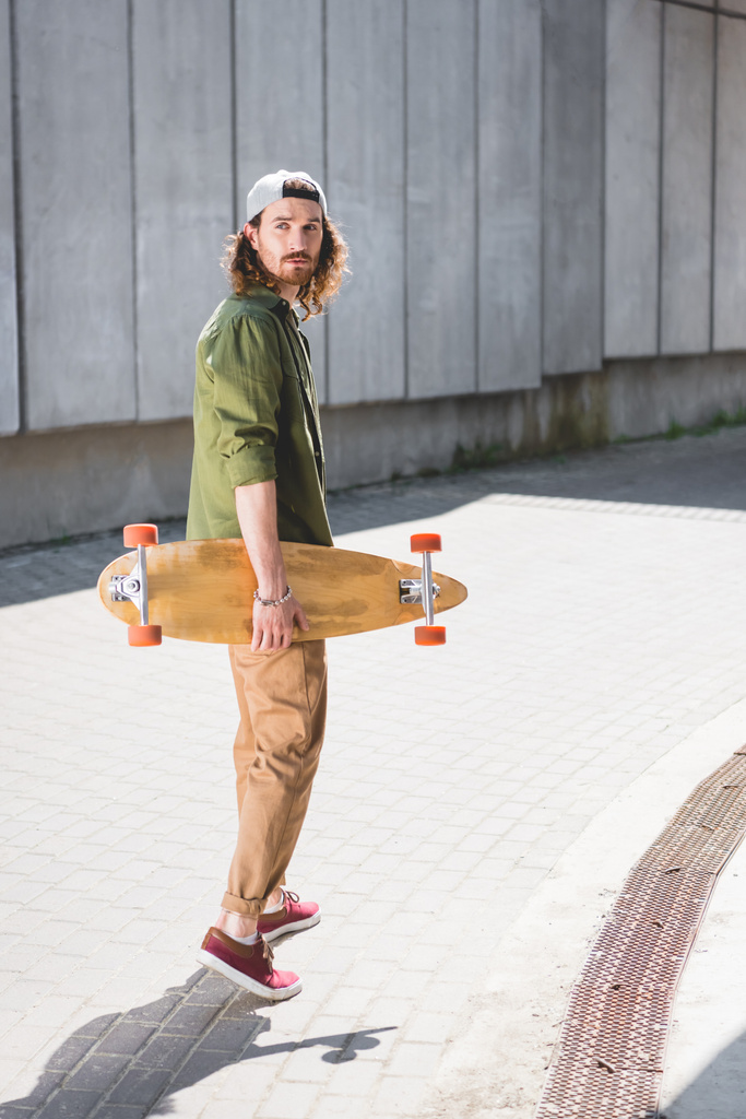 handsome man in casual wear standing near concentrate wall with skateboard in hand, looking away - Photo, Image