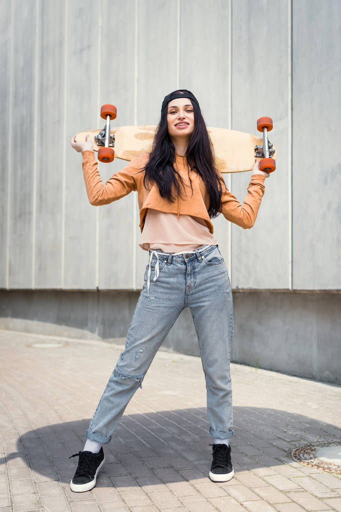 Fröhliche Frau in Freizeitkleidung steht an der Wand und hält Skateboard hinter dem Rücken - Foto, Bild