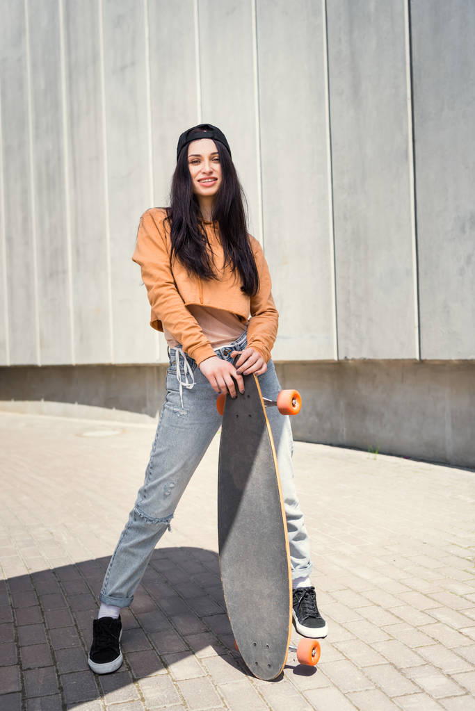 brunette woman in casual wear standing near concentrate wall with skateboard in hands - Photo, Image