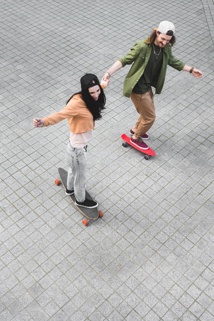 vista ad alto angolo di coppia felice che si tiene per mano, sorridente e cavalcando su skateboard
 - Foto, immagini