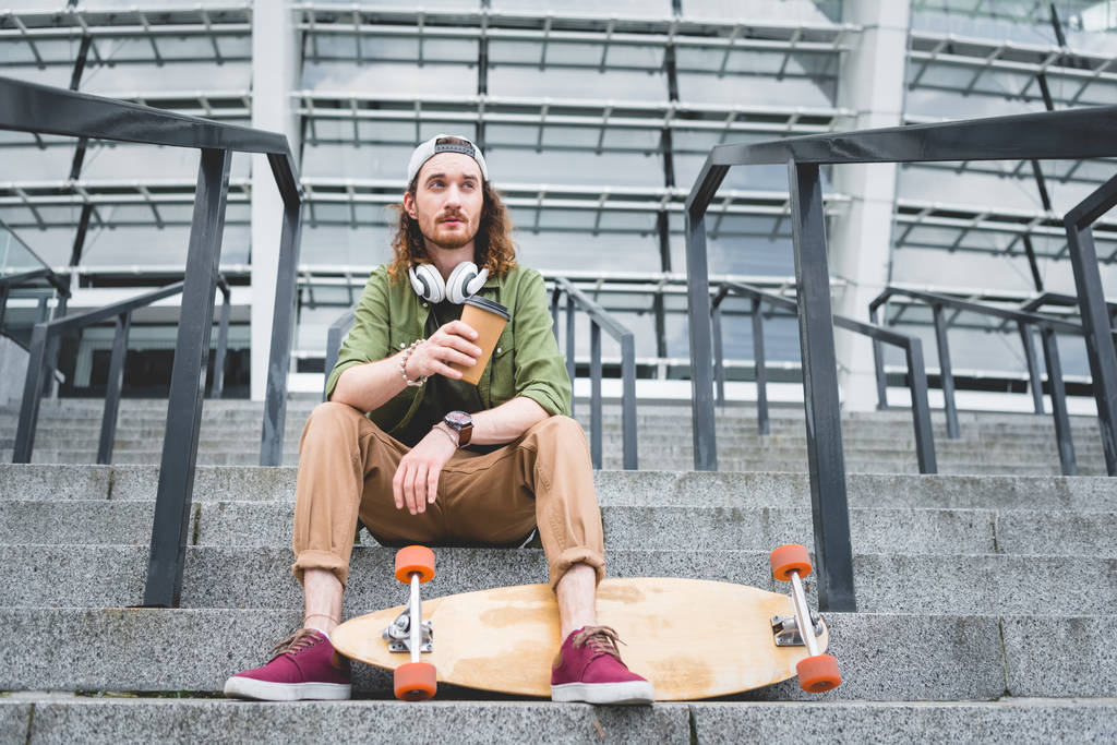 lage hoek weergave van kalm man met papier Cup in de hand op zoek weg, zittend op de trap in de buurt van skateboard - Foto, afbeelding