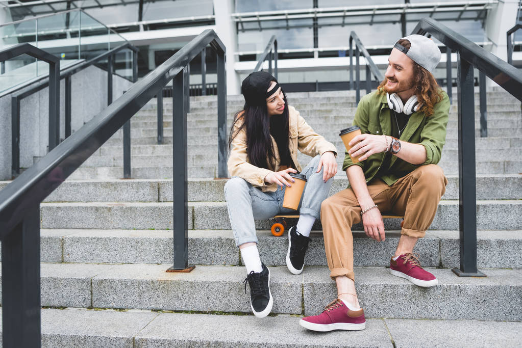 nízký úhel pohledu na ženu a muže sedící na skateboard s papírovým šálkem v ruce, který se dívá na sebe - Fotografie, Obrázek